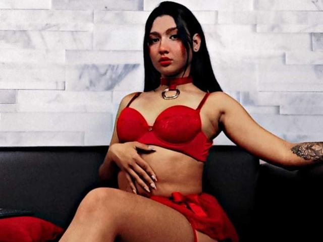 PauletteRoy with long black hair, red bra, and matching red skirt, wearing a red choker, sitting on a black couch, hand on her belly, tattoo on right arm.
