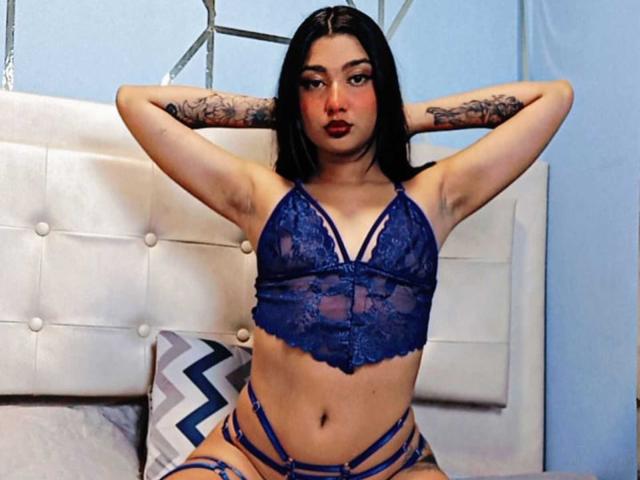 PauletteRoy with black hair, blue lace bra, and matching strappy lingerie, arms raised, showing tattoos, and a confident, sexy expression, sitting on a bed with a white headboard.
