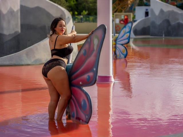 Lolita, com corpo avantajado e bumbum grande, vestindo lingerie de renda preta, segurando um adereço de borboleta colorida, em pé em água vermelha, fazendo um sinal de positivo com o polegar, sorrindo, com cabelo escuro e pele bronzeada..