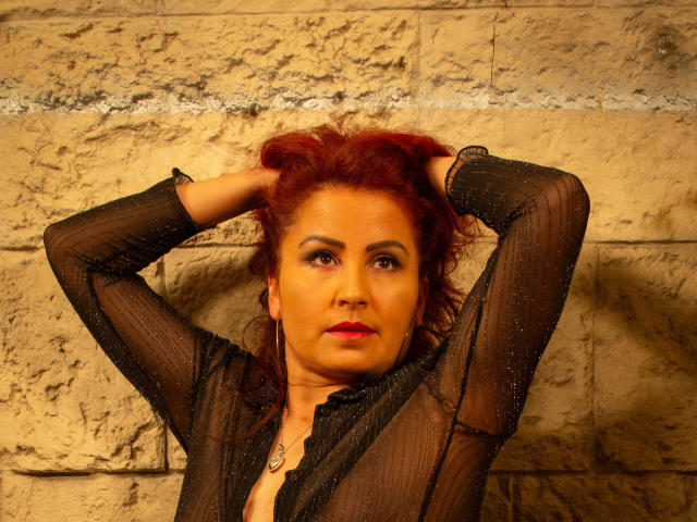 SussyFantasy a les cheveux roux, elle porte un haut noir transparent et a les mains dans les cheveux, mettant en valeur sa poitrine et son rouge à lèvres rouge, avec un mur de pierre rugueuse en arrière-plan..