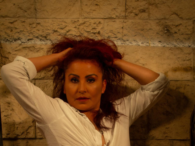SussyFantasy has red hair, hands in it, wearing a white shirt, looking serious, with a tan, slightly rough-skinned face, and a small nose. She's in a dimly lit room.