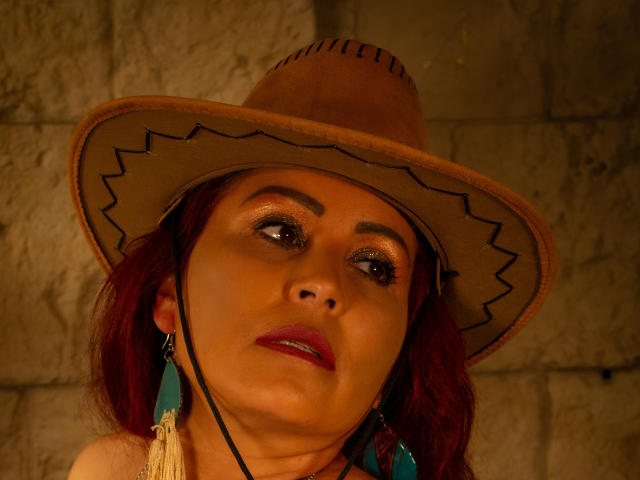 SussyFantasy avec des cheveux roux, un chapeau de cowboy marron, des boucles d'oreilles turquoise et du rouge à lèvres rouge, regardant vers le haut avec des yeux scintillants, une bouche légèrement ouverte et un cordon noir près de son visage..