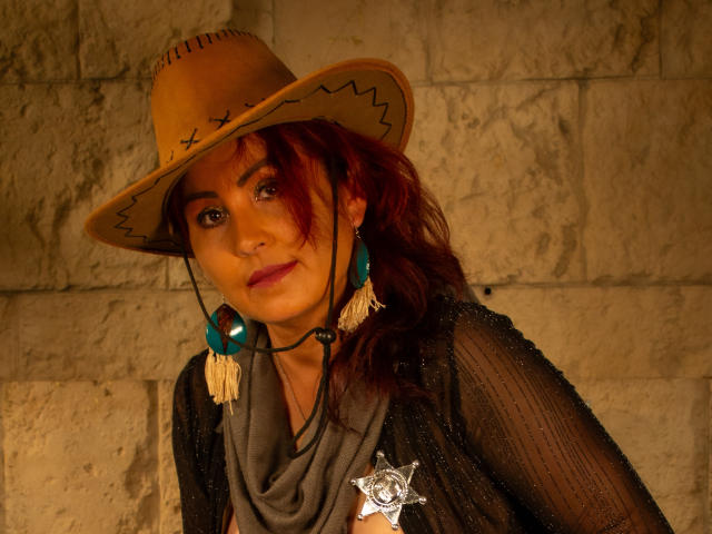 SussyFantasy with red hair, wearing a cowboy hat, teal earrings, black sheer top, and a sheriff's star, has a cute smile and a sexy, confident look.
