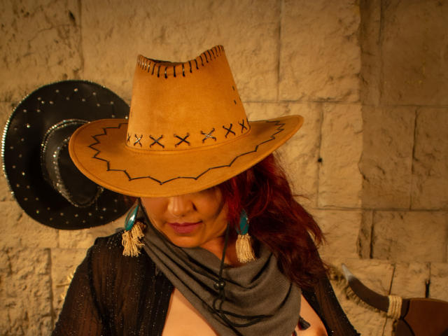 SussyFantasy com cabelo ruivo, usando um chapéu de cowboy bege com "X"s pretos, cachecol cinza e blusa preta transparente, mostrando os seios, e franjas turquesa, em uma luz quente..