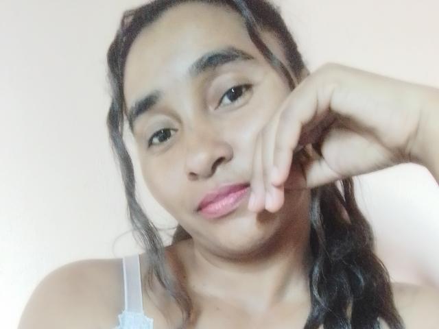 Ramonah with dark hair in a braid, brown skin, pink lipstick, and a slight pout, touching her cheek with a finger. She's wearing a white strap.