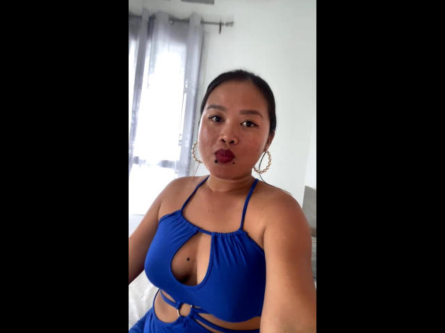 NellahHottie mit mittelgroßer Brust, trägt ein blaues Kleid mit Ausschnitten, hat geschwollene Lippen und trägt Ohrringe, macht ein Selfie in einem hellen Raum..