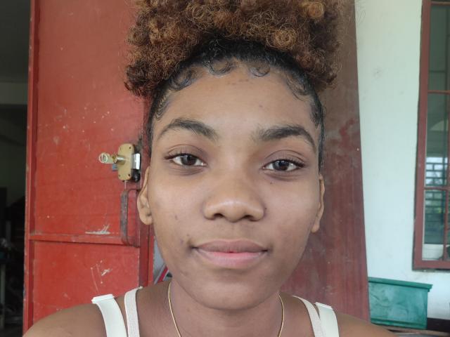 LisaK has curly brown hair, brown eyes, and a slight smile. She wears a white tank top and a thin gold necklace, with a small beauty mark under her right eye.