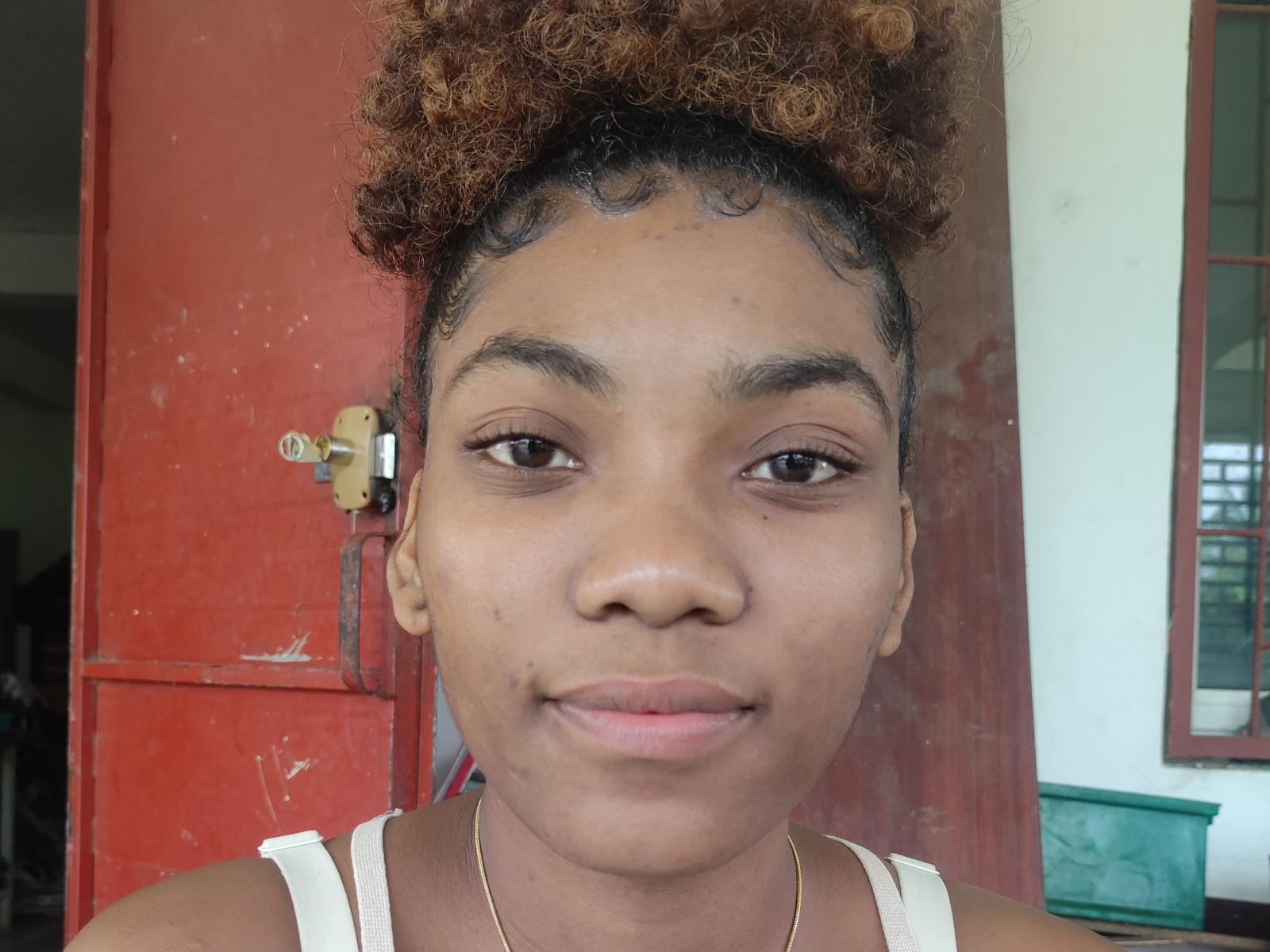 LisaK has curly brown hair, brown eyes, and a slight smile. She wears a white tank top and a thin gold necklace, with a small beauty mark under her right eye.