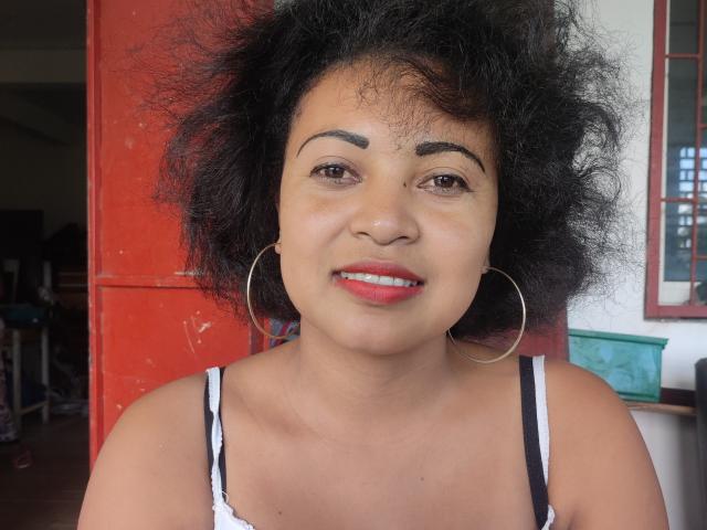 Delinah hat lockiges, schwarzes Haar, braune Augen und trägt roten Lippenstift mit großen Ohrringen. Sie lächelt und hat kleine, runde Brüste und eine niedliche Nase. Sie trägt ein weißes Tanktop..