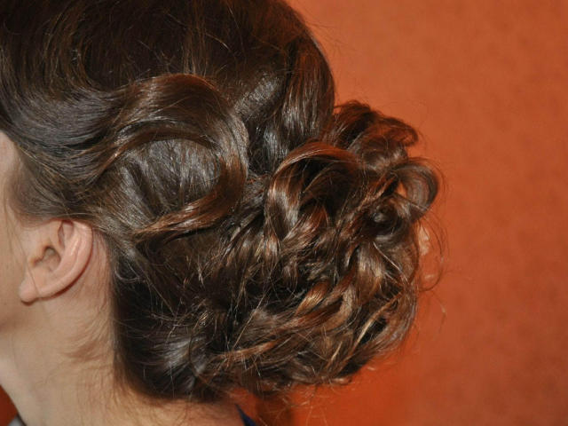 Keiri's bruine, krullende opgestoken kapsel is een strak, elegant knotje met krullende lokken, dat haar glanzende, golvende haar laat zien tegen een oranje achtergrond..