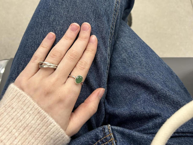 Keiris Hand mit einem grünen Edelsteinring und einem silbernen Armband am Finger, ruht auf ihrer blauen Jeans, sie trägt einen beigen Pullover und man sieht ihre rosa Nägel..