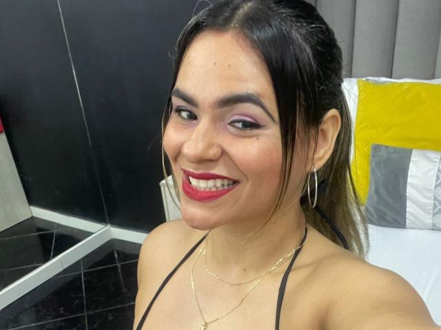 BrianaPreston has brown hair, red lipstick, purple eyeshadow, hoop earrings, and a black halter top, smiling with her tits barely visible, wearing a gold necklace.
