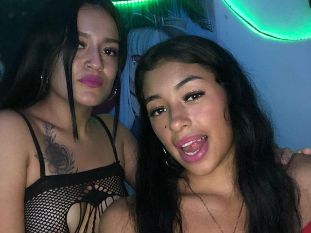 KratosXLadies with long black hair, pink lipstick, and black fishnet top showing small tits, stands beside KratosXLadies with brown hair, pink lipstick, and tongue out, in a selfie.