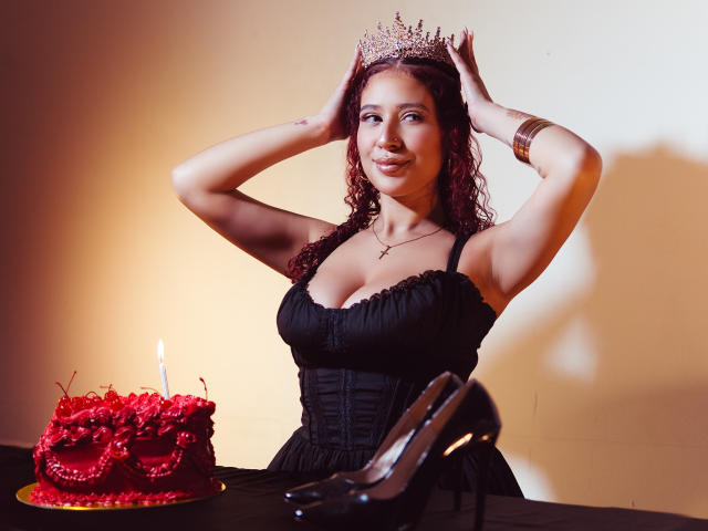 ValeryScot with curly red hair, wearing a black dress, tiara, and bracelet, smiling, hands on head, black high heels, red cake with candle, and a small cross necklace.