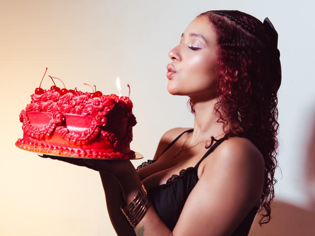 ValeryScot with curly red hair, black dress, and purple eyeshadow, kissing a red, rose-decorated cake with a candle, holding it in her hands. She's got a tattoo on her arm.