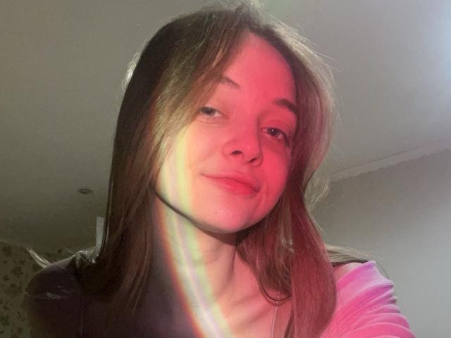 MayxFlower's face with pinkish-red light, brown hair, slight smile, rainbow reflection on skin, wearing pink top, light from above, casual selfie, soft light, subtle shadow on face.