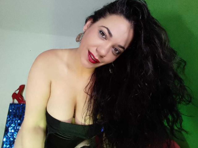 YolittS has long black curly hair, red lipstick, and is wearing a green strapless top showing her big boobs. She's smiling with red earrings in a green-lit setting.