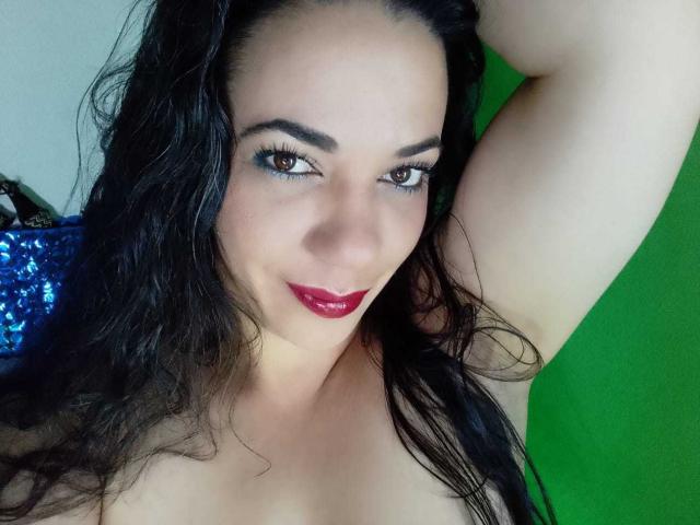 YolittS has dark hair, red lipstick, and blue eyeliner, with her arm raised, showing her bare shoulder and a green background. Boobs partially visible. Smiling, eyes looking at camera.