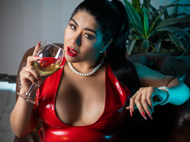 Khloe Lawson, vestindo um vestido de látex vermelho, um colar de pérolas e batom vermelho, segurando uma taça de vinho, sentada em um sofá de couro marrom, com seus seios grandes e unhas vermelhas visíveis, cabelo escuro em um rabo de cavalo..