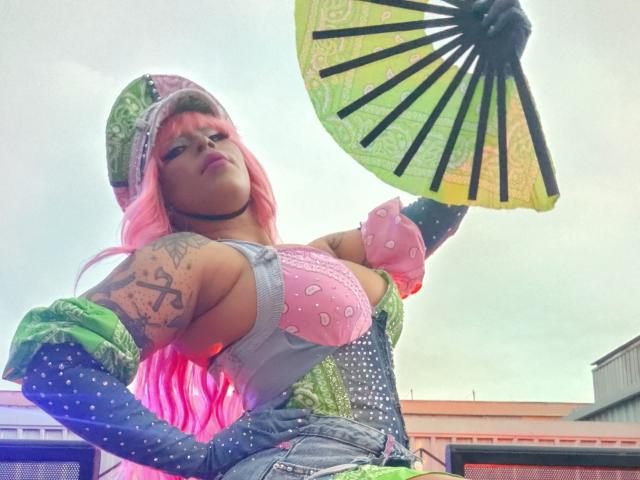 Chili with pink hair and tattoos is wearing a colorful outfit, holding a green fan, and looking up confidently.