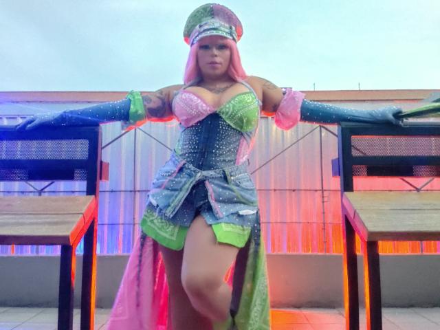 Chili with pink hair wearing a sparkly green and pink outfit and hat, leaning on black table, showing thigh, legs spread.