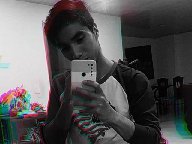 LucasSmith with short dark hair, wearing a ragged shirt, holding a white smartphone, taking a selfie, with colorful 3D-style effects. His face is slightly downcast, eyes focused.