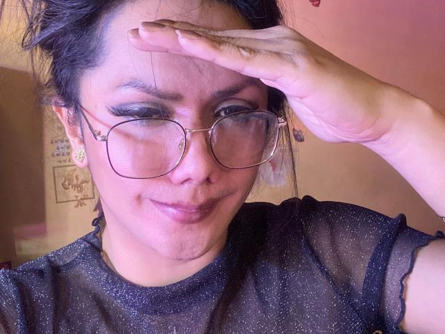 Asiantastic-hot with glasses, black sheer top, and a subtle smile, touches forehead, showing off her cute nose and shiny lips. Her hand is up, covering part of her face.
