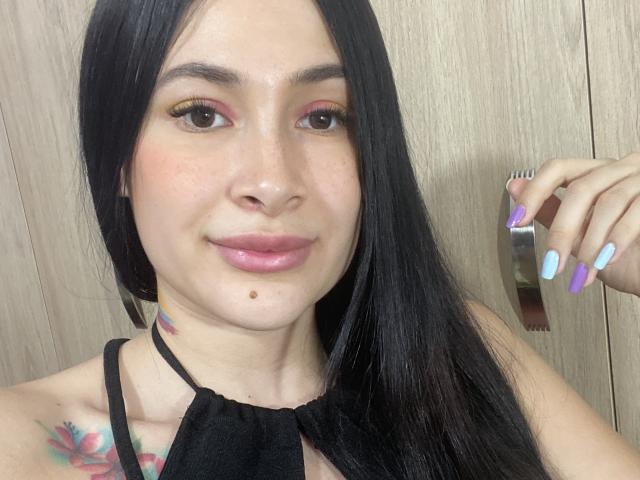 CherryLoux with long black hair, pink eyeshadow, glossy pink lips, small moles, colorful nail polish, black halter top, tattoo on left shoulder, holding a metal object.