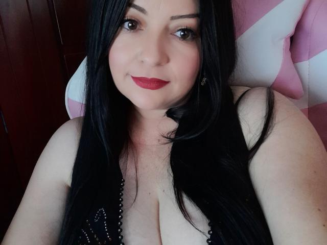 NatalyHami with long black hair, red lipstick, and black lace top, showing her large boobs, sitting on a pink and white chair, smiling at the camera.