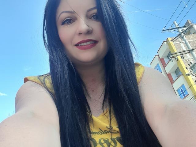 NatalyHami has long black hair, pale skin, and red lipstick, wearing a yellow sleeveless top, smiling with red lips, and a nose ring, taking a selfie outdoors.