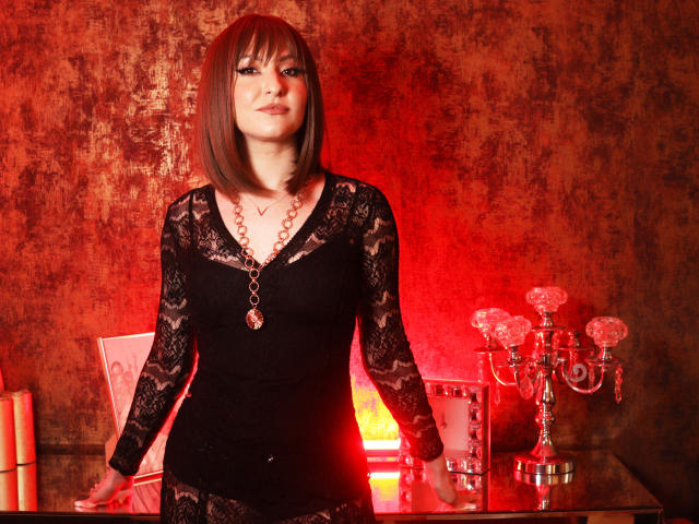 GoddessAlma with brown bob, wearing black lace dress, necklace, and red light. Her tits are small, she's standing with arms on glass table, holding crystal candelabrum.