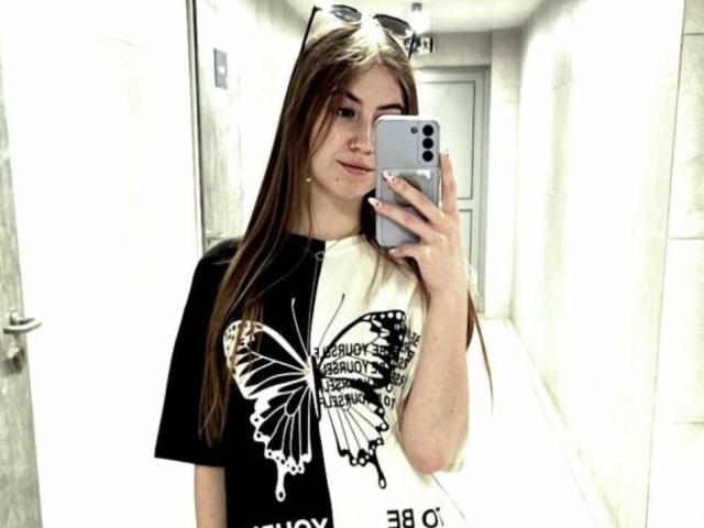 MeganCandy with long brown hair, wearing a black and white butterfly shirt, holding a silver iPhone, standing in a bright, narrow hallway, smiling at the mirror.