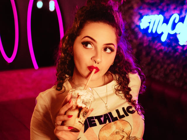 CharlotteAdam with curly brown hair, red lipstick, drinking from a cup with a straw, wearing a white "Metallica" shirt, looking up with a pouty expression, neon pink and blue lights.