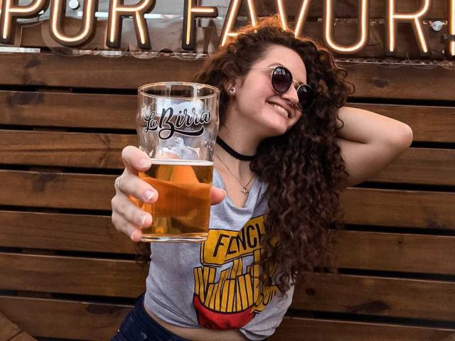 CharlotteAdam with curly brown hair, wearing sunglasses and a choker, holding a beer glass, smiling, wearing a "Fence Varmint" shirt, and blue jeans.