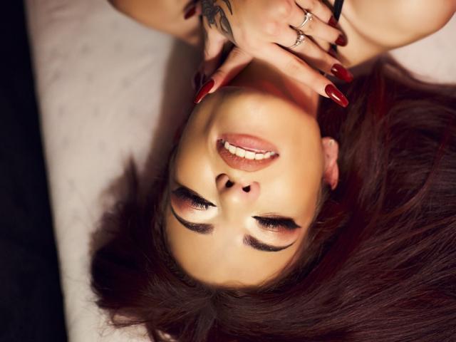 RinaQueenie's face upside down, dark red nails, red lipstick, tattoo on hand, smiling with eyes closed, wearing black top, white background, red nails covering her mouth.