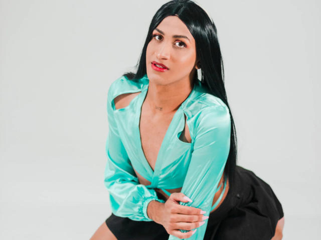 BarbaraPerri69 with long black hair, wearing a shiny turquoise blouse with cutouts, black shorts, and a neutral expression, sitting with hands in front, looking up.