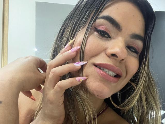 BrianaPreston has long, blonde-tipped hair, pink eyeshadow, and pink lipstick. She's smiling with her hand near her face, showing pink and yellow nail art. Her tits are slightly visible.