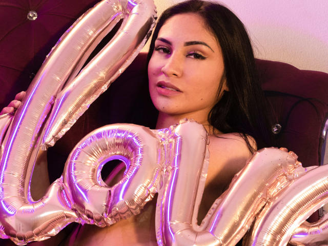 LanaBox with shiny pink "20" balloons, holding them near her chest. She has long dark hair, light skin, and is wearing pink eyeliner. Her lips are slightly parted.