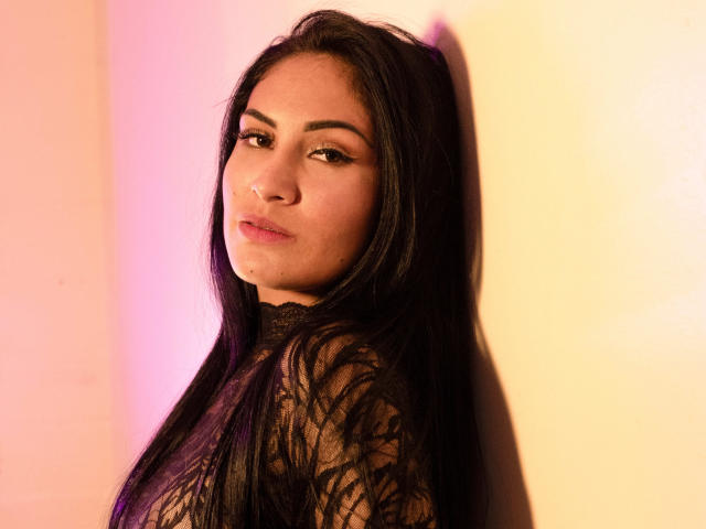 LanaBox with long black hair, wearing a lace top, looking over her shoulder, pink and yellow lighting on her face, subtle makeup, slightly pouty expression, confident, sexy.