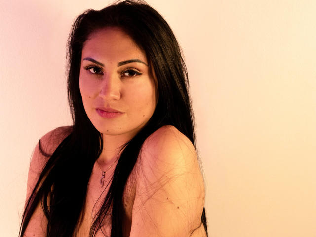 LanaBox with long black hair, light skin, and pink lipstick, looking at the camera with a slight smile, wearing a necklace, and showing her bare shoulders and small tits.