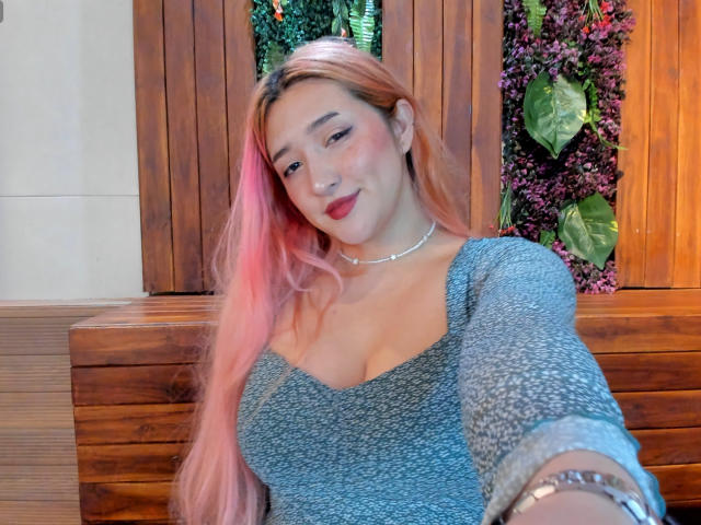 IsabellaClown with pink hair, wearing a grey, long-sleeve top, showing her tits, smiling, red lipstick, silver necklace, wooden background, and green leaf on the right.