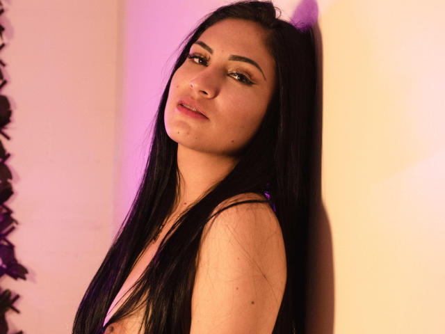 LanaBox with long black hair, slightly tilted head, and subtle makeup, looking to the side with a relaxed expression, standing against a yellow wall with pink lighting.