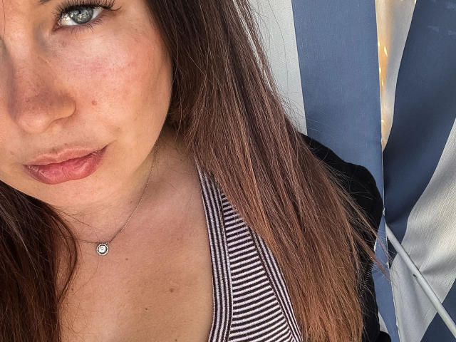 Shyla, séduisante avec sa peau claire, ses cheveux bruns, ses yeux bleus et ses lèvres roses, porte un débardeur rayé qui dévoile sa poitrine et une petite chaîne avec un pendentif..