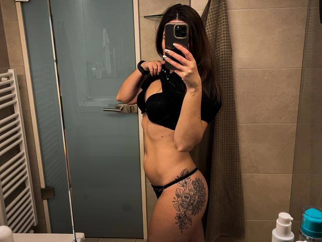 Shyla, attraente in reggiseno e slip neri, mostra un tatuaggio sulla coscia destra, tiene un telefono in un selfie allo specchio, con un addome piatto e un seno di medie dimensioni..