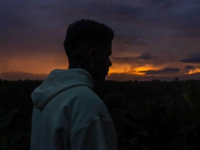 Silhouetted photo of AlexaSandoval in a white hoodie, back to the camera, with dark, short hair, facing a sunset with orange and purple clouds in the sky.