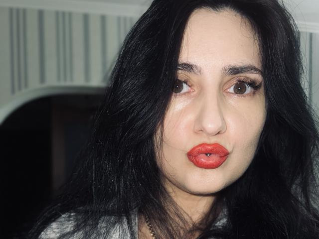 JennaEasy has pale skin, dark wavy hair, brown eyes, and bright red lipstick with slightly parted full lips. Her nose is small, and she's wearing a gray shirt.