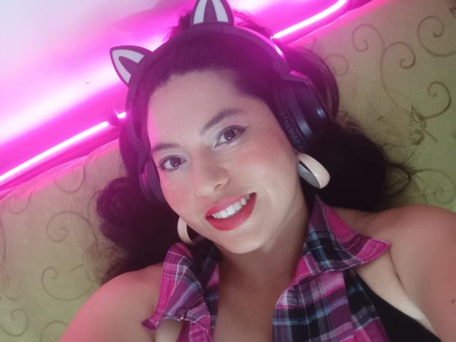 ElizabetCati, com uma tiara de orelhas de gato, sorrindo, vestindo uma camisa xadrez rosa, fones de ouvido brancos grandes, luzes neon rosa e batom vermelho, tem seios médios e pele marrom clara..