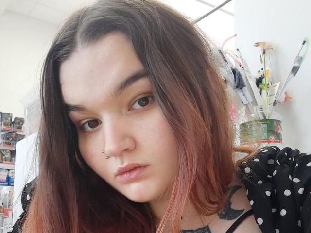 Eva Berg tem pele clara, olhos castanhos, um piercing no nariz e cabelo castanho-avermelhado, vestindo uma blusa preta com bolinhas, com uma expressão séria, em um quarto claro..