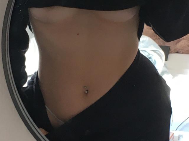 Mirror selfie of SweetHottBabe69 showing a flat stomach with a belly button piercing, black clothes partially pulled down, and exposed tan lines from small, rounded tits.