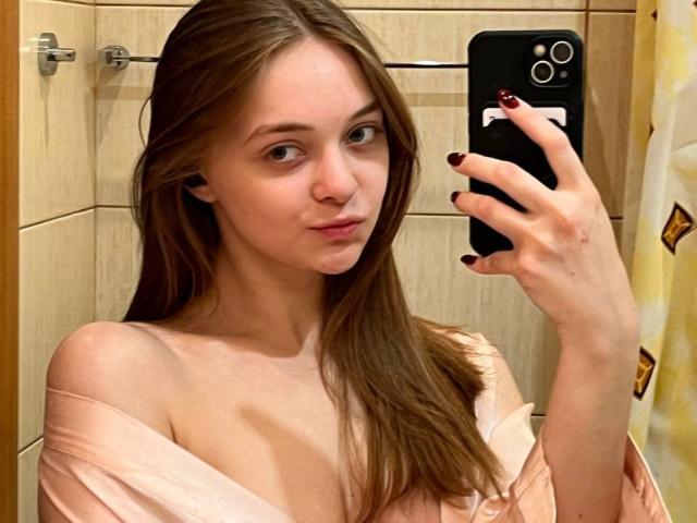 MayxFlower tirando uma selfie com um celular em um banheiro, mostrando sua pele clara, cabelo castanho e blusa rosa com um ombro de fora, expondo seus pequenos seios. Suas unhas estão pintadas de cores escuras..